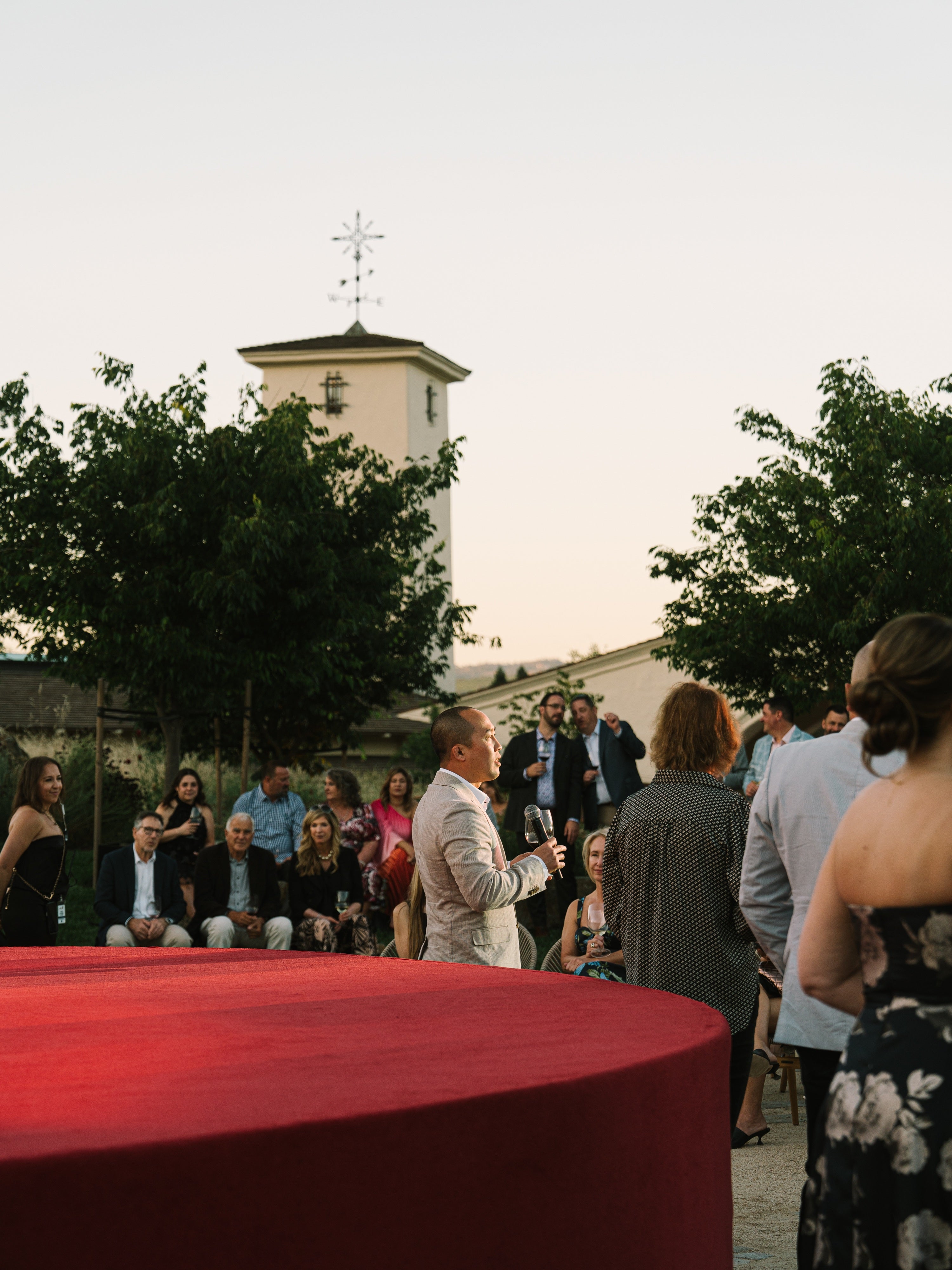An outdoor evening event scene with a crowd of guests gathered around a table covered in a red cloth, with a man speaking into a microphone. Trees and a building with a tower are visible in the background under a pale sky.