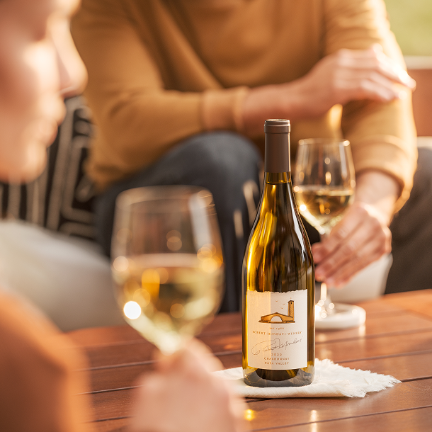 A bottle of Robert Mondavi Winery Chardonnay on a table with people holding wine glasses.