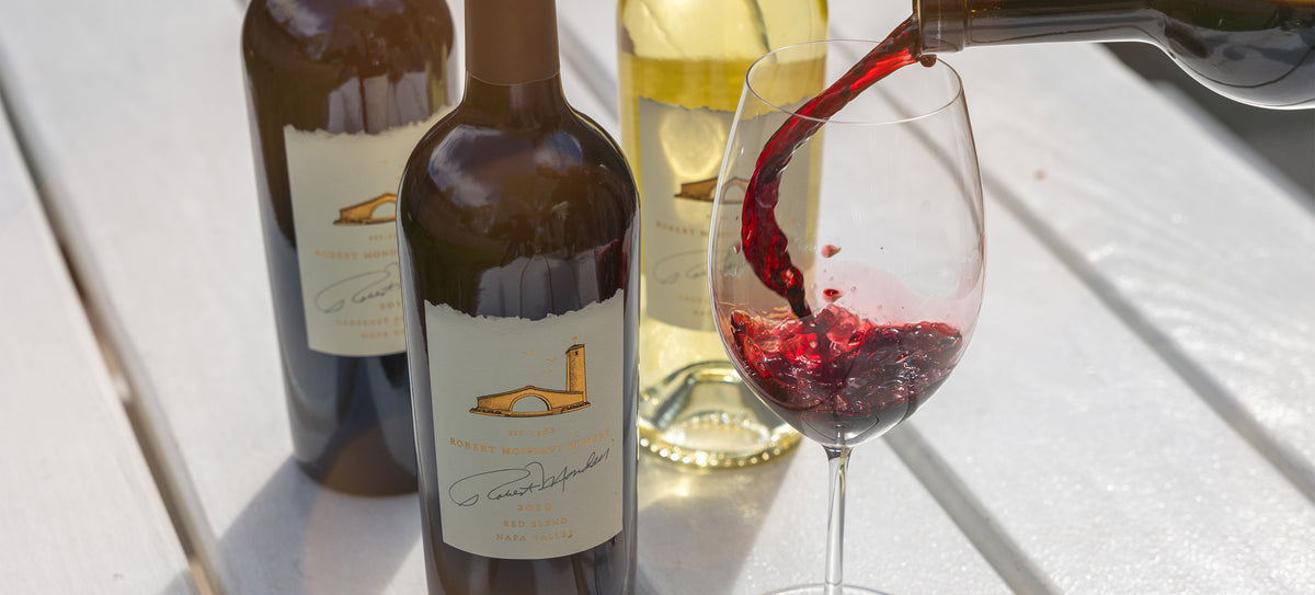 3 Robert Mondavi wines sit on a white table with a red wine being poured into a glass.