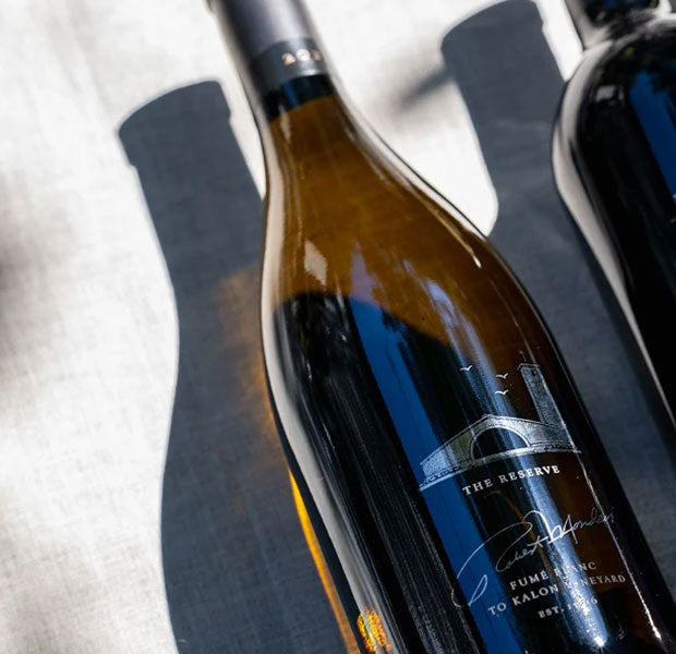 A close-up of a wine bottle labeled 'The Reserve' from Robert Mondavi Winery, resting on a textured light gray surface with strong natural shadows.