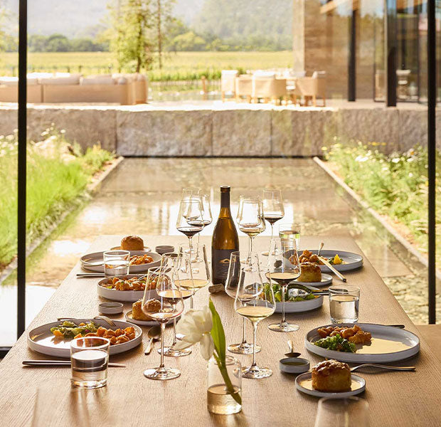 A long dining table set with plates, wine glasses, a bottle, and food in a bright, modern dining room with large windows overlooking a landscaped outdoor area.