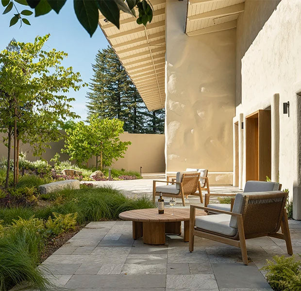 Modern outdoor patio furniture set including two wooden armchairs with light gray cushions, a round wooden coffee table, and a wicker side table, arranged on a stone patio next to a stucco building with a landscaped garden.