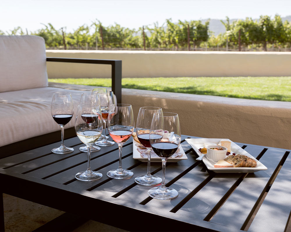 4 glasses of wine and small snacks, sit on a table. The background is the lawn and the vineyards.